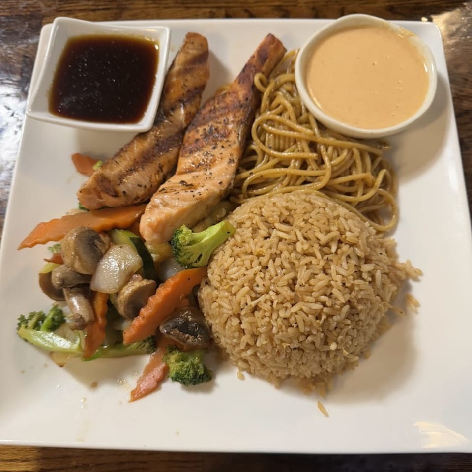 Grilled salmon with rice, noodles, mixed vegetables, and two dipping sauces on a white plate.