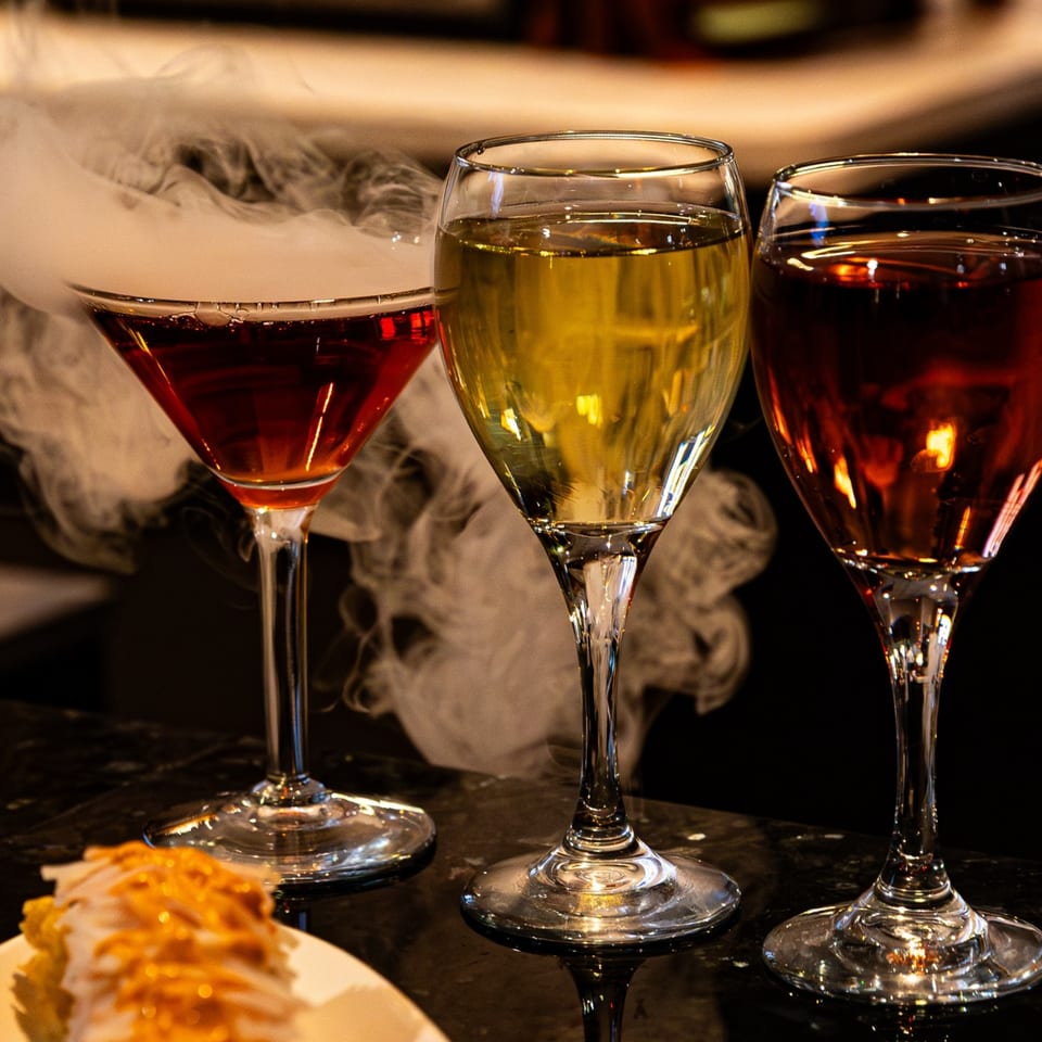 Three drinks, one smoking, on a dark bar with a plate of food in the foreground.